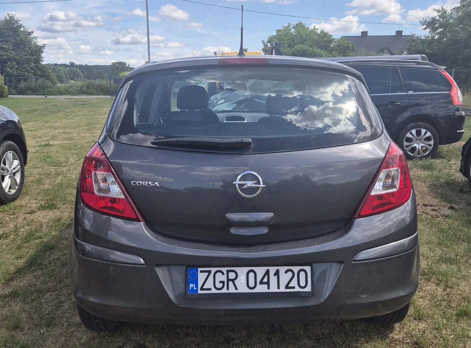 Tylny widok szarego samochodu marki Opel Corsa zaparkowanego na trawie. Na klapie bagażnika widnieje napis 'CORSA' oraz logo marki Opel. Samochód posiada polską tablicę rejestracyjną o numerze 'ZGR 04120'. W tle widoczne jest niebo z chmurami, zieleń oraz fragment innego, ciemnego samochodu po prawej stronie.