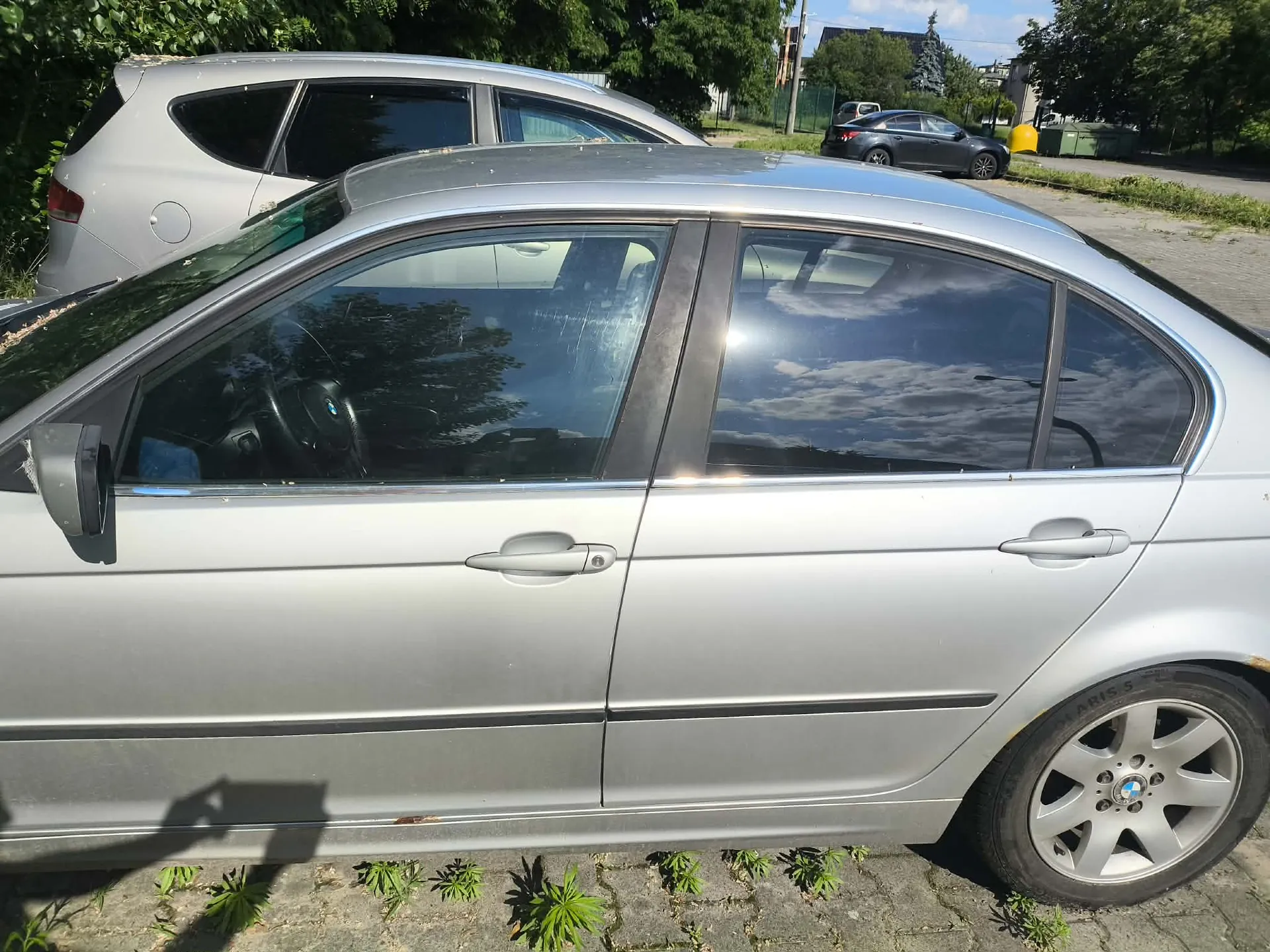 Srebrny samochód osobowy w widoku z boku, zaparkowany na kostce brukowej. W tle widoczny jest inny srebrny samochód oraz ciemny pojazd zaparkowany dalej na ulicy, a także drzewa i fragmenty zabudowań.