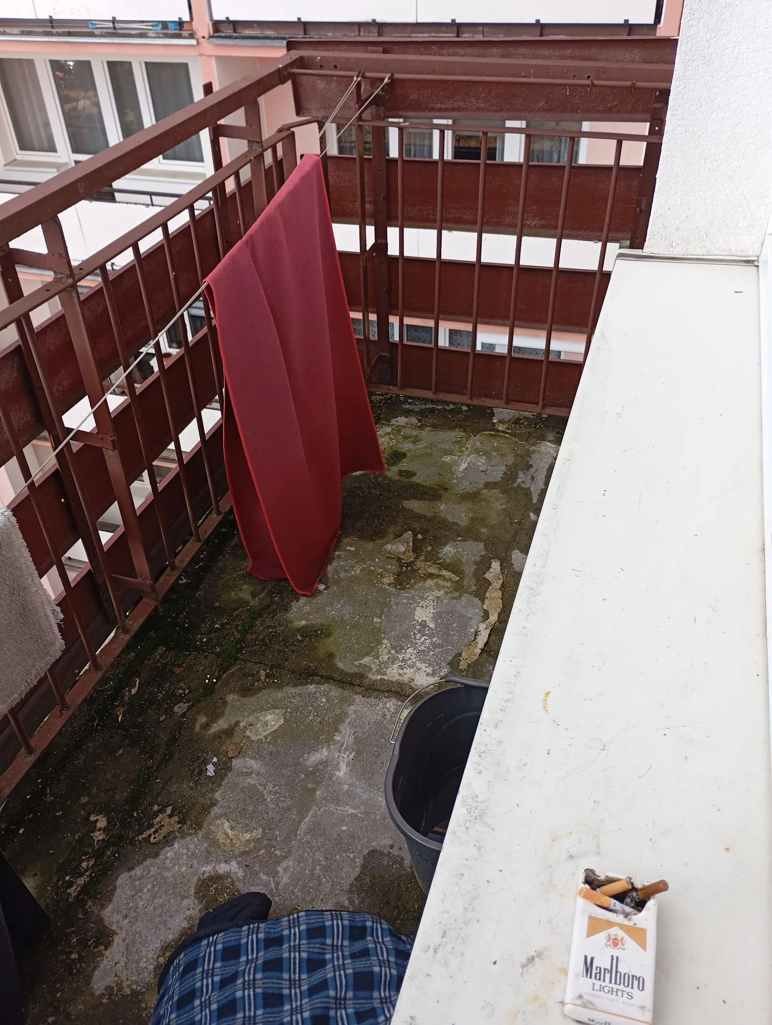 Widok z góry na betonowy balkon z brudną, zaplamioną podłogą. Po lewej stronie znajduje się brązowa metalowa barierka, na której wisi czerwony koc. W prawym dolnym rogu widoczna jest biała krawędź murku, na którym leży paczka papierosów marki Marlboro Lights. Obok murku stoi czarny plastikowy pojemnik, a na podłodze leży niebieska koszula w kratkę.