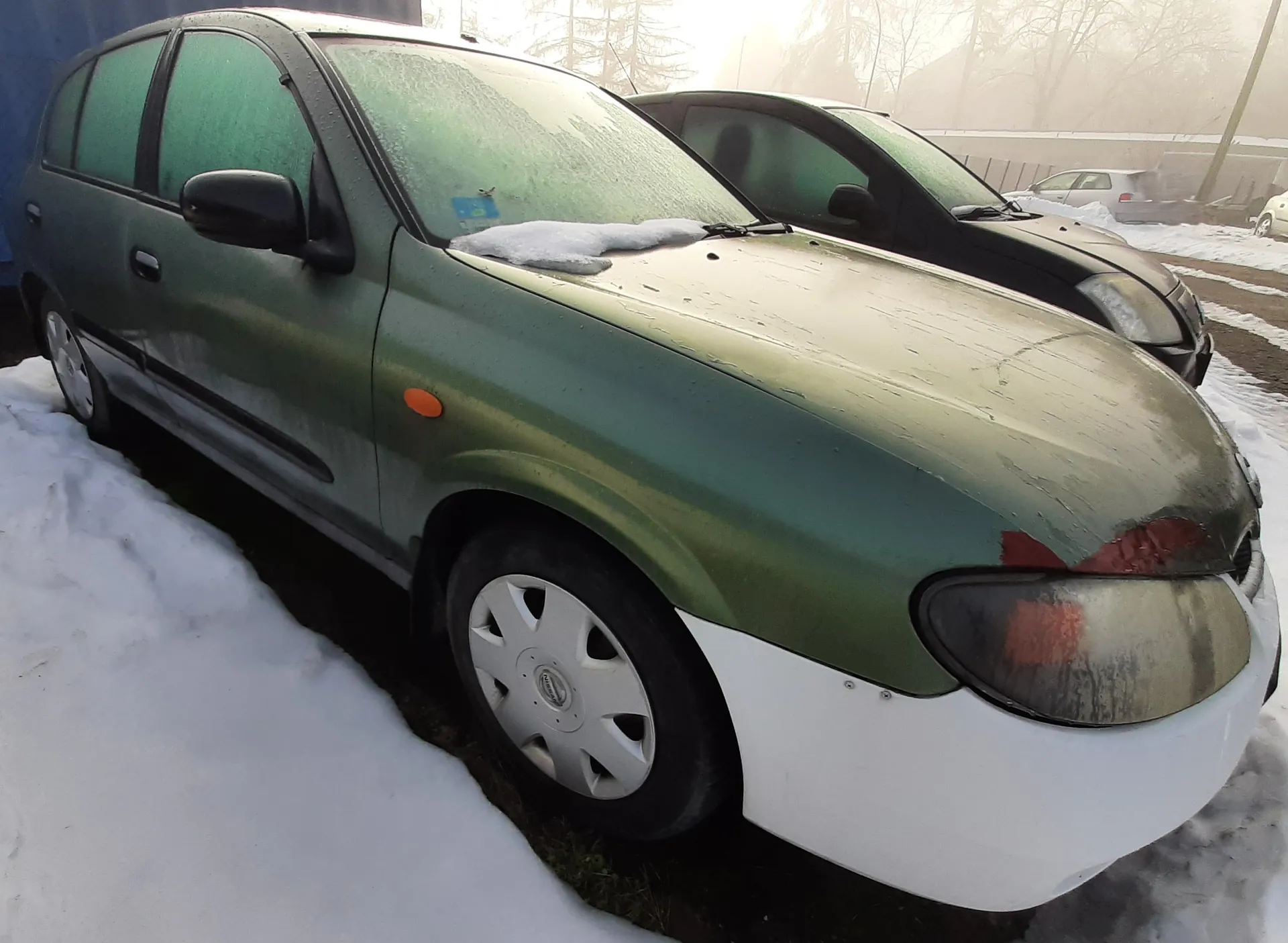Zielony samochód osobowy z białym zderzakiem przednim zaparkowany na śniegu. Na masce widoczna warstwa śniegu, a na przedniej szybie niebieski naklejony znaczek. Samochód ma pomarańczowy odblask na boku i srebrne felgi. W tle drugi czarny samochód, drzewa bez liści, budynki oraz śnieżna droga. Słońce świeci w tle, tworząc mglistą atmosferę.