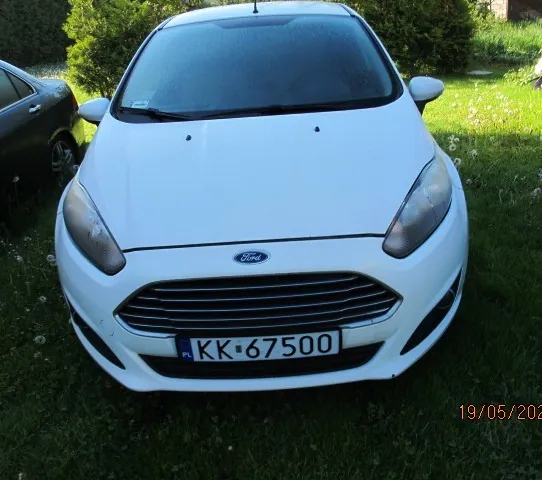 Biały samochód marki Ford z przodu, z tablicą rejestracyjną KK-67500. Po lewej stronie częściowo widoczny czarny samochód. Tło stanowi trawa, krzewy i fragment domu. W prawym dolnym rogu widoczna data 19/05/2020.
