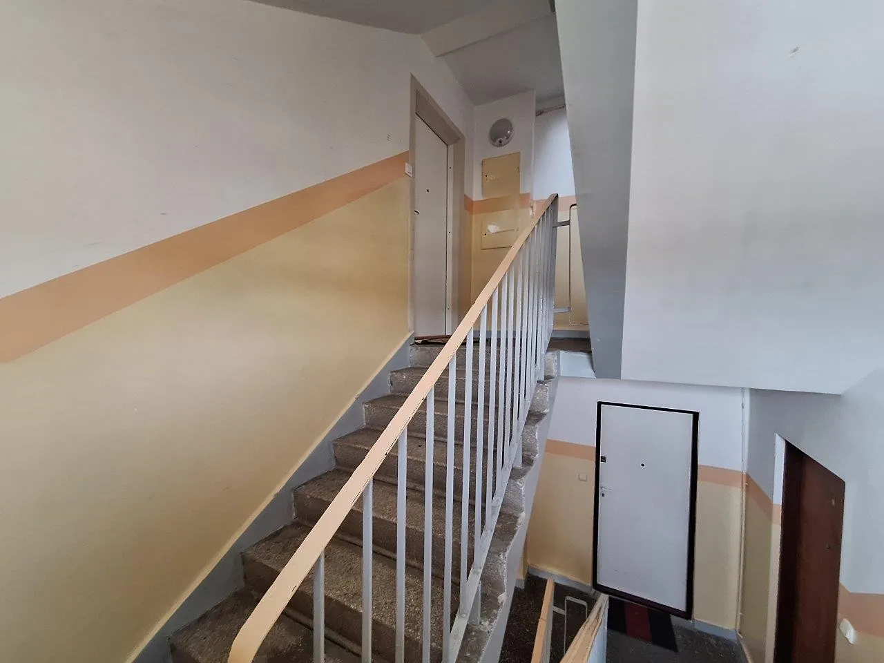 Klatka schodowa wewnątrz budynku. Betonowe schody prowadzą w górę, zabezpieczone białą, metalową balustradą z pomarańczową poręczą. Ściany są pomalowane na dwa kolory: jasny beż w dolnej części i biały w górnej, rozdzielone poziomym, pomarańczowym pasem. Na górnym podestcie widoczne są zamknięte drzwi, a na dolnym poziomie po prawej stronie znajdują się białe drzwi wejściowe oraz ciemnobrązowe drzwi obok nich.
