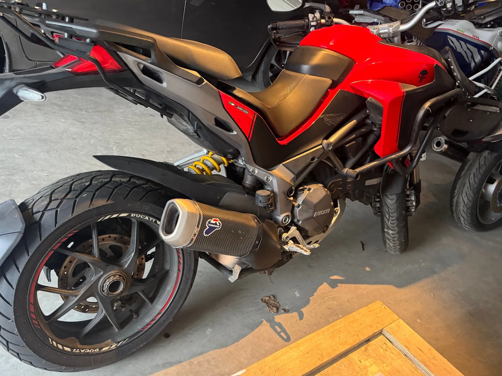 Zbliżenie na prawą stronę czerwono-czarnego motocykla marki Ducati. Widoczne są detale konstrukcyjne: tylne koło z czarną felgą i czerwonym paskiem na oponie, wydech z karbonowym wykończeniem, żółta sprężyna amortyzatora oraz czerwony bak. Motocykl stoi w garażu na betonowej podłodze.