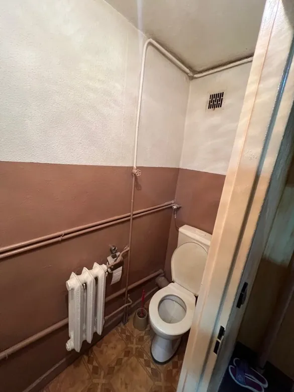 Łazienka z białą toaletą poziomo umieszczoną, białym umywalką, ścianami w kolorze brązowym i białym, widocznymi miedzianymi rurami, białym grzejnikiem, prostokątnym wentylatorem na ścianie oraz podłogą wyłożoną płytkami w geometryczny wzór. W prawym górnym rogu obrazu znajdują się fioletowe klapki.