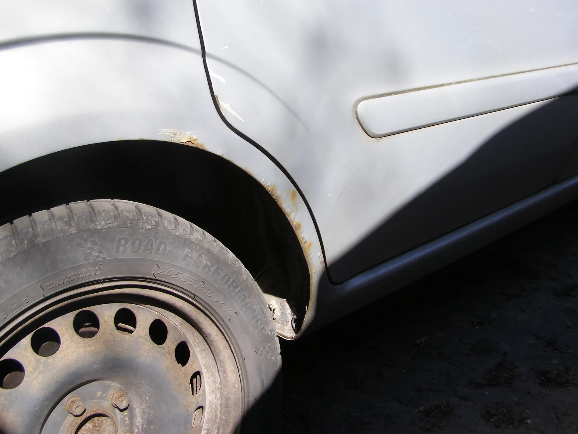 Zbliżenie na przednią część białego samochodu, konkretnie na koło i nadkole. Widoczna jest opona z napisem "ROAD PERFORMANCE" oraz oznaczeniem rozmiaru "205/55 R16 94V". Felga ma metalowy kolor z otworami i jest lekko zabrudzona. Wokół nadkola widoczne są ślady rdzy. Tło stanowi ciemna, brudna powierzchnia podłogi.