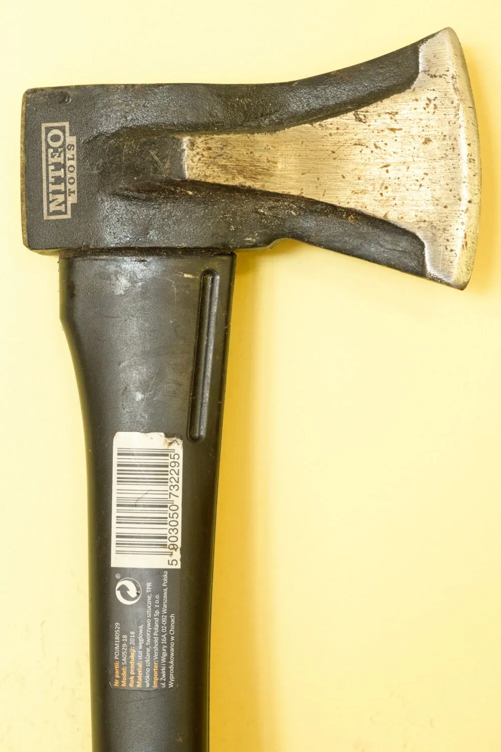 Zdjęcie siekiery z czarnym trzonkiem i metalową głowicą na jasnym tle. Na głowicy widnieje napis 'NITEO TOOLS'. Na trzonku znajduje się biała naklejka z kodem kreskowym oraz tekstem w języku polskim, zawierającym informacje o modelu, roku produkcji i producencie.