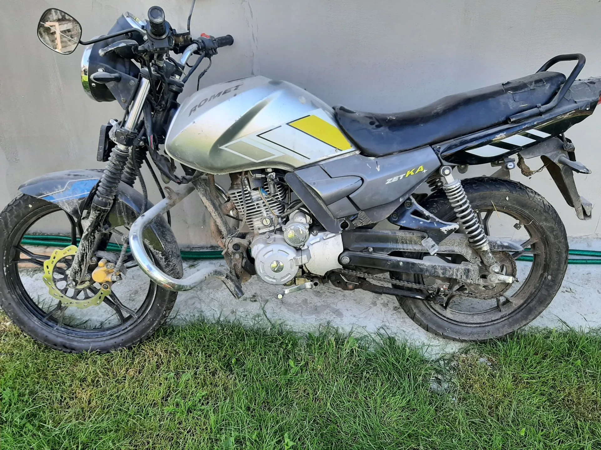 Srebrny motocykl marki Romet, model Zetka, zaparkowany na trawie przy jasnej ścianie. Pojazd ma czarną kanapę, żółte akcenty na baku oraz widoczne zabrudzenia na kołach i dolnych częściach silnika.