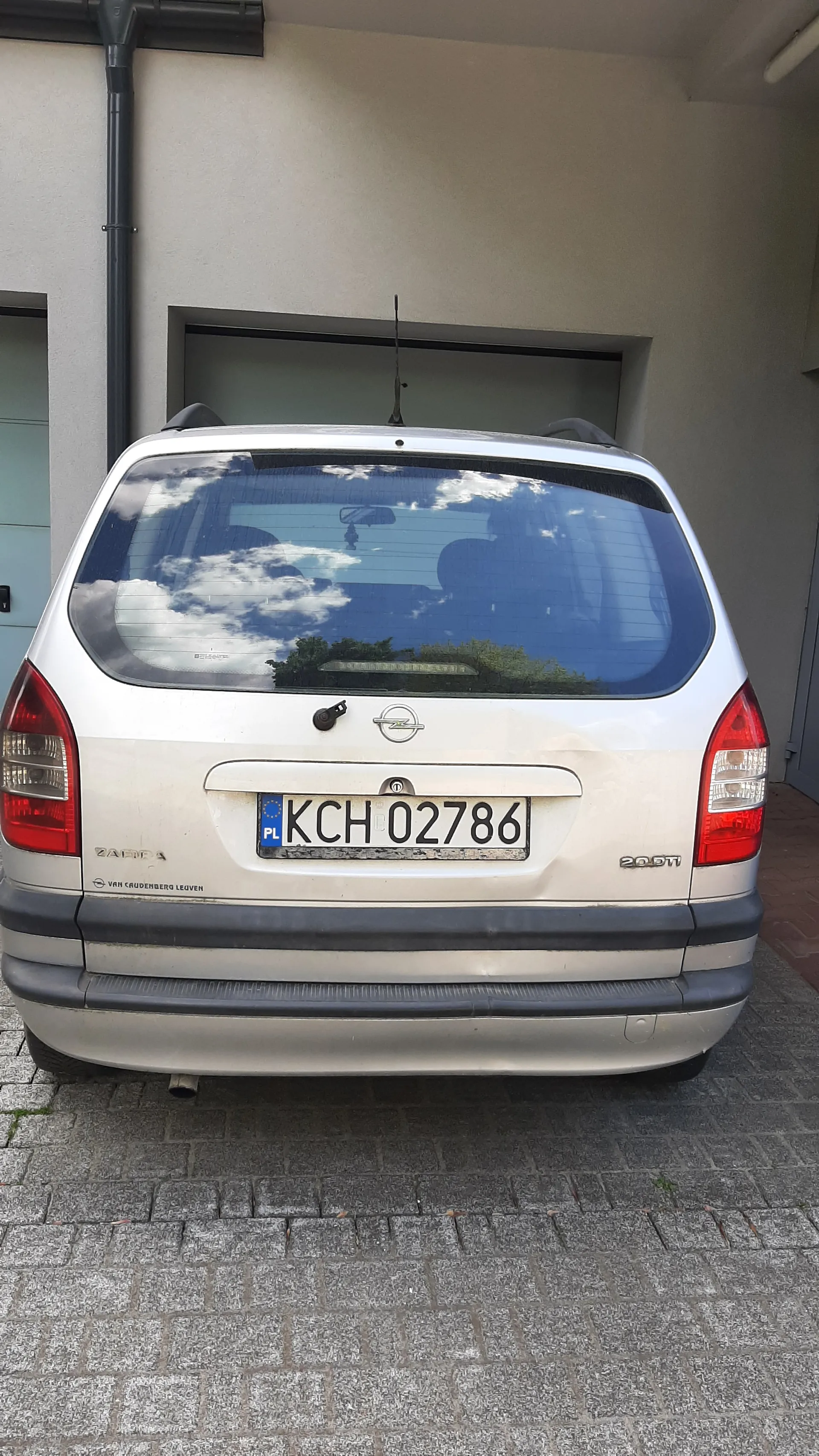 Tył srebrnego samochodu marki Opel Zafira z tablicą rejestracyjną KCH 02786. Na tablicy widoczny niebieski pas z oznaczeniem PL oraz flagą UE. Samochód zaparkowany na brukowanej powierzchni obok budynku o beżowej ścianie z oknem. W tylnej szybie odbijają się chmury i drzewa. Na bagażniku widoczne napisy '20DT' oraz 'ZAFIRA'.