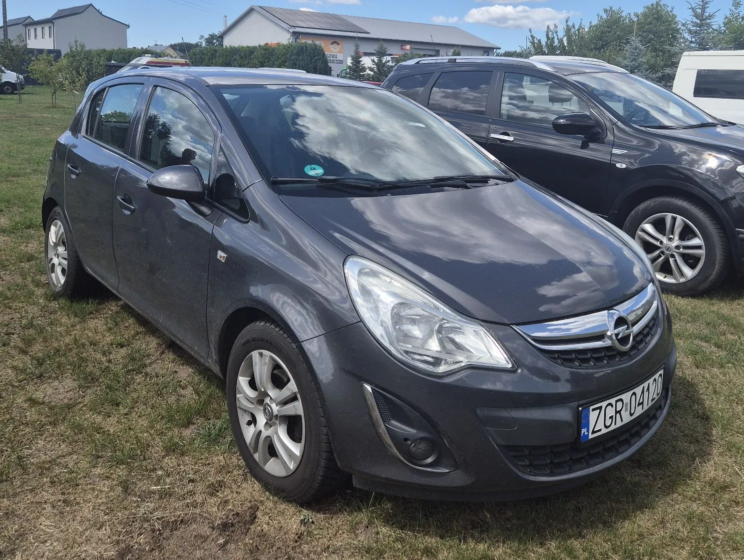 Szary samochód osobowy marki Opel zaparkowany na trawiastym terenie. Pojazd jest ustawiony pod kątem, widoczny jest przód oraz lewy bok auta. Na przednim zderzaku znajduje się polska tablica rejestracyjna o numerze ZGR 04120. W tle widoczne są inne zaparkowane samochody, niskie budynki mieszkalne oraz drzewa pod jasnym niebem z lekkimi chmurami.