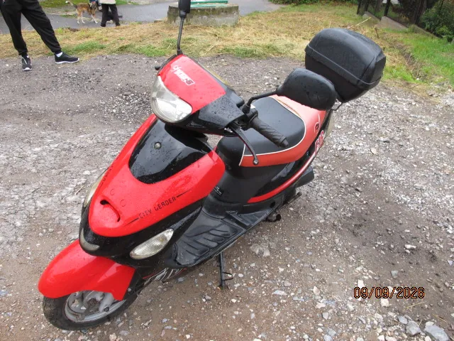 Czerwono-czarny skuter marki Yamaha z modelem 'CITY LEADER' napisanym na przedniej owiewce. Pojazd posiada zamontowany czarny kufer z tyłu oraz lusterka boczne. Skuter stoi zaparkowany na szutrowym podłożu. W tle widoczne są nogi osoby w czarnych spodniach i butach oraz fragment trawnika.
