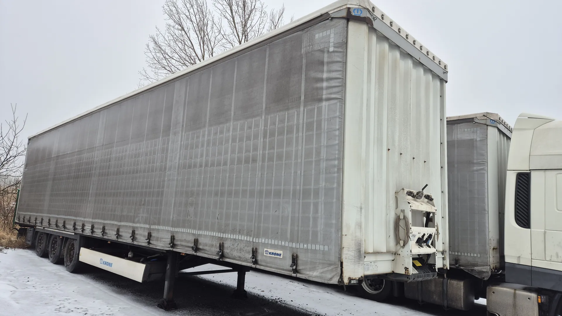 Duży przyczepa ciężarowa z szarą tkaniną pokrywającą ładunek, biała kabina ciężarówki po prawej stronie. Na przyczepie widoczne logo KRONE w niebieskim kolorze. Tło: nagie drzewa, szare niebo, śnieg na ziemi. Przyczepa stoi na nawierzchni częściowo pokrytej śniegiem.