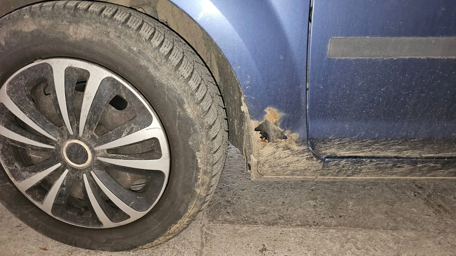 Zbliżenie na przednie lewe koło niebieskiego samochodu. Nadbrzeże nad kołem jest zabrudzone błotem i posiada wyraźne uszkodzenie w postaci dziury spowodowanej korozją blachy.