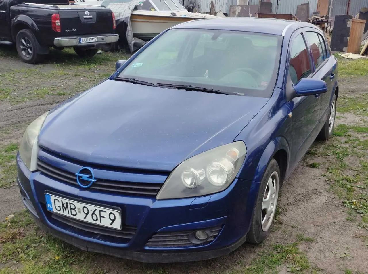 Niebieski samochód marki Opel z tablicą rejestracyjną GMB 96F9 (PL) na pierwszym planie. W tle czarny pickup Nissan z tablicą CCH 09Y7 oraz łódź przykryta tarpem. Samochody stoją na nierównym terenie z trawą i żwirem, w tle widoczne są budynki i materiały budowlane.
