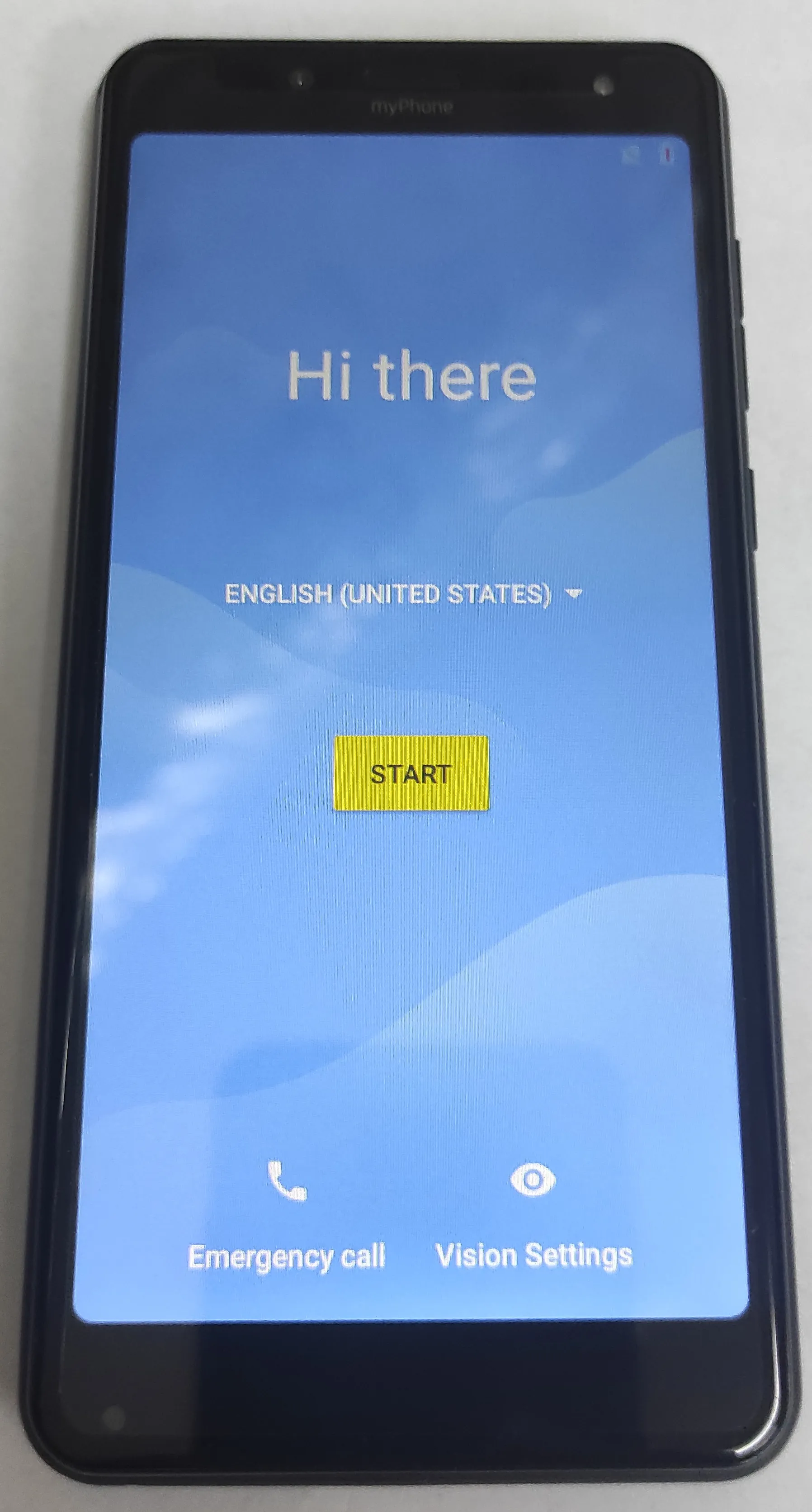 Smartfon marki myPhone z włączonym ekranem. Na niebieskim tle widnieje napis 'Hi there', rozwijane menu wyboru języka z zaznaczoną opcją 'ENGLISH (UNITED STATES)' oraz żółty przycisk z napisem 'START'. Na dole ekranu znajdują się dwie ikony z podpisami: 'Emergency call' oraz 'Vision Settings'.