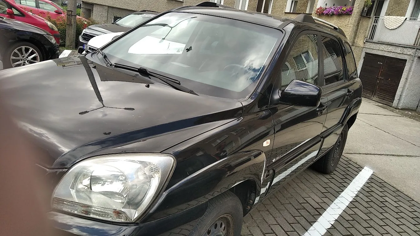 Czarny samochód osobowy typu kombi zaparkowany na kostce brukowej. Widoczny jest przód i prawy bok pojazdu, w tym reflektor, maska z odbiciem nieba oraz lusterko boczne. W tle widoczne są inne zaparkowane samochody oraz fragmenty budynków mieszkalnych z balkonami.