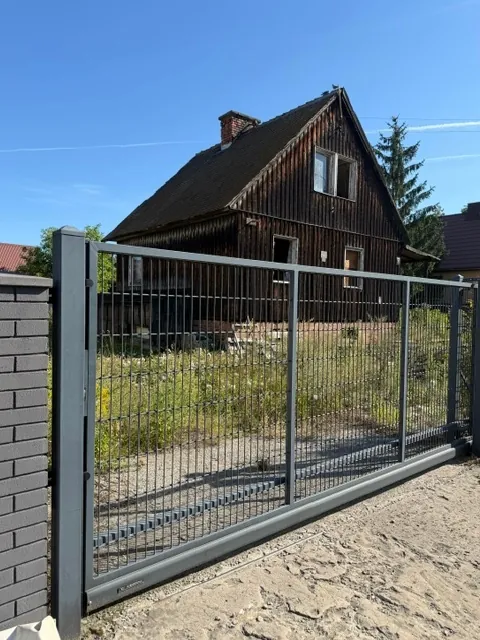 Zdjęcie przedstawiające szary, metalowy płot z siatki, zamontowany na betonowym słupku i przylegający do ciemnoszarego murka z cegieł. W tle znajduje się stary, drewniany dom z ciemnobrązową elewacją i dwuspadowym dachem, otoczony dziką roślinnością i trawą. Niebo jest bezchmurne i błękitne.