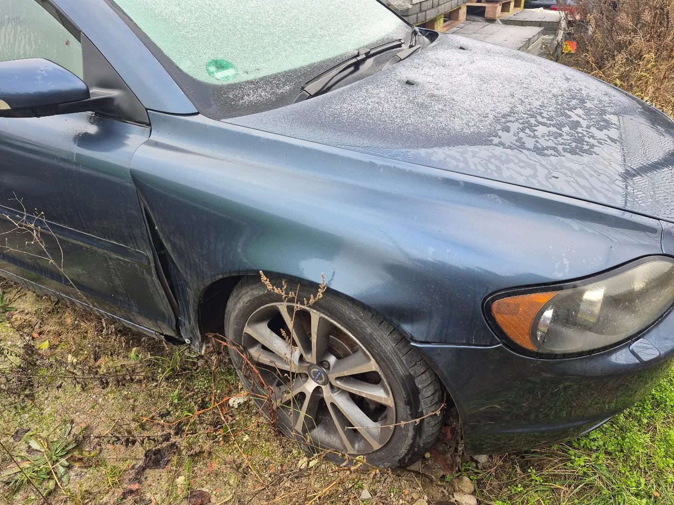 Ciemnoniebieski samochód osobowy zaparkowany na trawiastym i błotnistym terenie. Widoczna przód i lewa strona pojazdu. Na masce i przedniej szybie znajduje się biały nalot, prawdopodobnie śnieg lub sól. Przednie lewe koło z aluminiową felgą, wokół sucha roślinność. W tle fragmenty drewnianych palet i zarośli. Na przedniej szybie zielony naklejony znaczek.