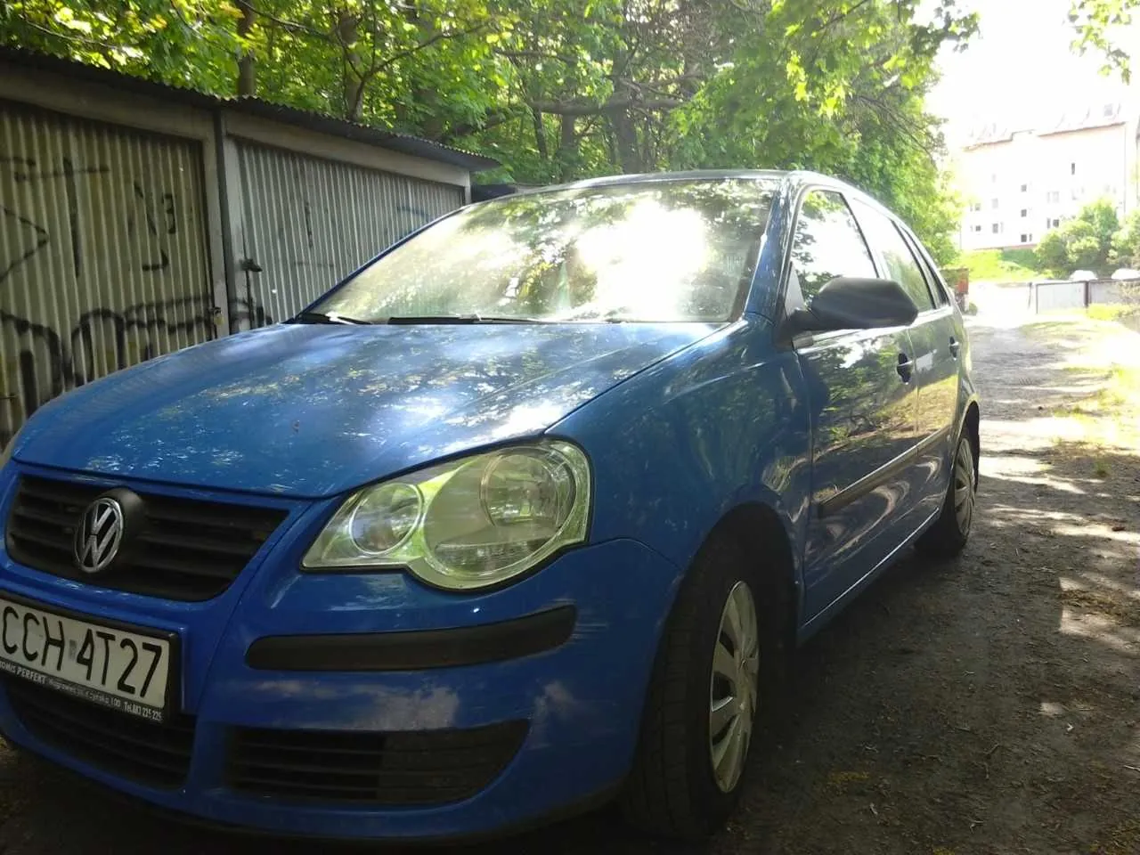 Niebieski samochód marki Volkswagen z tablicą rejestracyjną CCH 4T27 zaparkowany na drodze ziemnej. W tle metalowe garaże z graffiti, drzewa i budynek mieszkalny. Słońce oświetla scenę, tworząc cienie na ziemi.