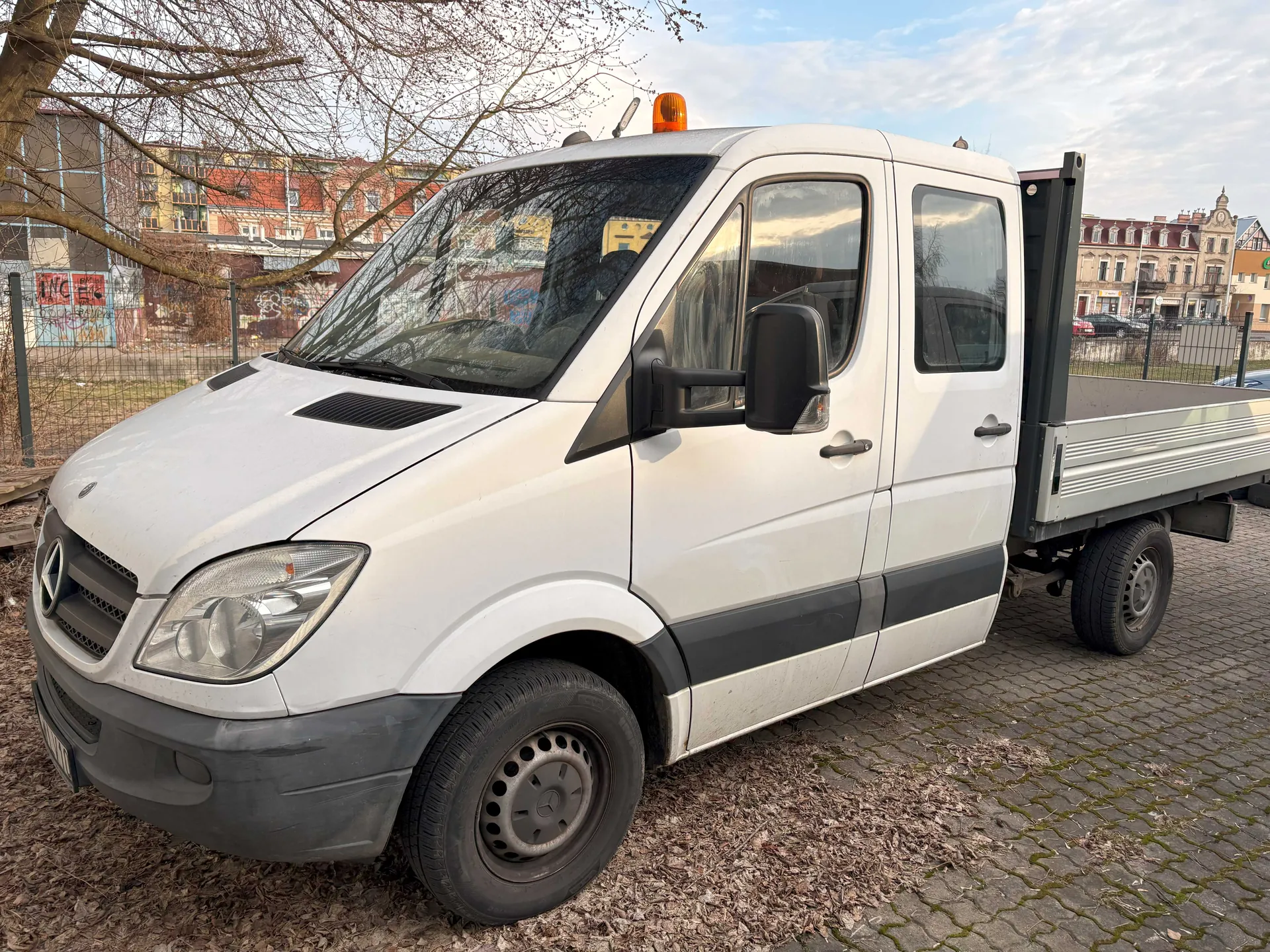 Biały samochód dostawczy marki Mercedes-Benz z platformą załadowczą, wyposażony w pomarańczowe światło ostrzegawcze na dachu. Pojazd stoi na brukowanej powierzchni, w tle widoczne są budynki mieszkalne z graffiti, drzewo bez liści oraz ogrodzenie. Na przedniej masce widoczny jest emblemat marki, a nad przednią szybą umieszczono lusterko wsteczne. Niebo jest częściowo zachmurzone.