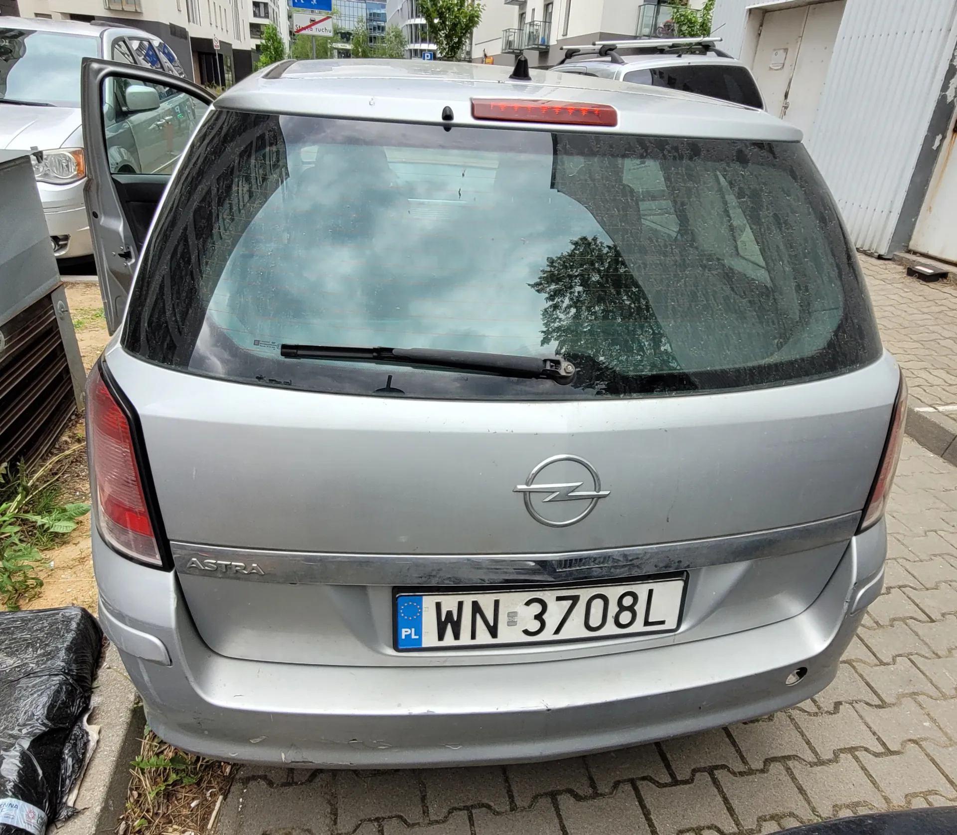 Z tyłu srebrny samochód Opel Astra z tablicą rejestracyjną WN 3708L (PL). Drzwi kierowcy są otwarte. W tle widoczne są inne pojazdy, budynki, drzewa oraz znak drogowy. Samochód stoi na brukowanej powierzchni, a na lewo od niego znajduje się czarna torba i metalowa konstrukcja.