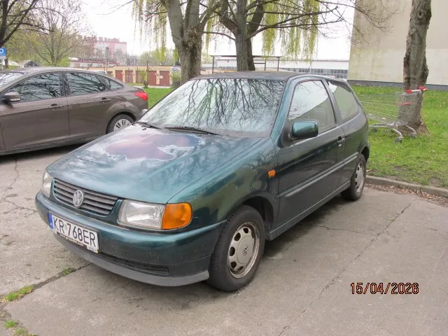 Zielony samochód osobowy marki Volkswagen z tablicą rejestracyjną KR 768ER zaparkowany na betonowej powierzchni. W tle widoczny brązowy samochód, drzewa, trawnik, budynki oraz datę 15/04/2026 w prawym dolnym rogu. Na pierwszym planie asfaltowa droga i fragmenty zieleni.