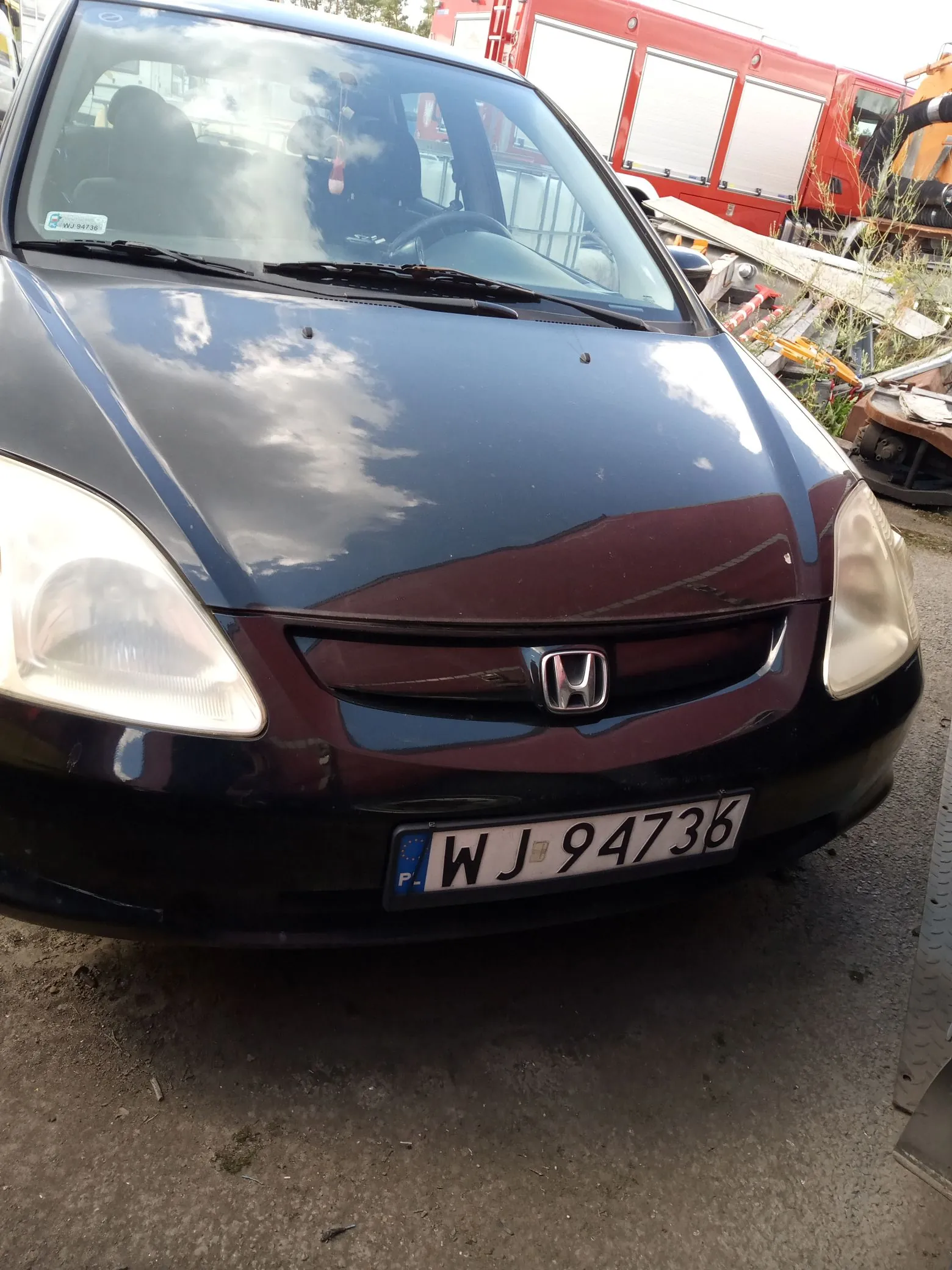 Przednia część ciemnogranatowego samochodu marki Honda. Widoczna jest maska z odbiciem chmur, reflektory, logo Hondy na grillu oraz polska tablica rejestracyjna o numerze WJ 94736. W tle widoczny jest czerwony pojazd ciężarowy oraz elementy złomowiska.