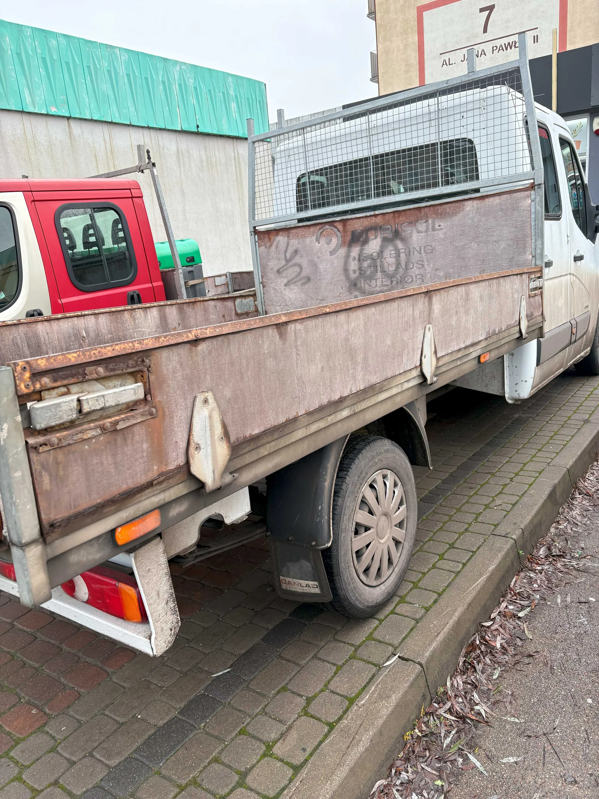 Zdjęcie przedstawia tył starszego, rdzewiejącego samochodu dostawczego z metalową siatką na platformie. Na bocznej ścianie platformy widoczny jest napis: BORISOL, ISOLERING, STILLADS, INTERIOR. W tle czerwony pojazd, budynek z zielonym dachem oraz tablica z napisem AL. JANA PAWŁA II i cyfrą 7. Samochód stoi na brukowanej nawierzchni, obok chodnika z suchymi liśćmi.
