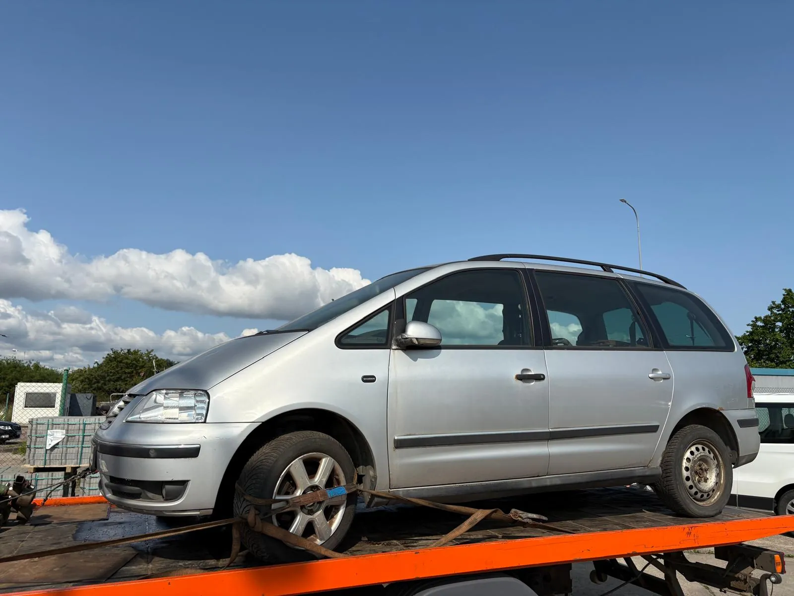 Srebrny minivan z czarnym dachowym bagażnikiem przewożony na pomarańczowej płycie holownika. Samochód jest przywiązany brązowymi paskami. Tło: błękitne niebo z białymi chmurami, zielone drzewa, biały samochód, latarnia uliczna oraz budynki. Widoczne są również elementy infrastruktury, takie jak siatka i stosy materiałów.