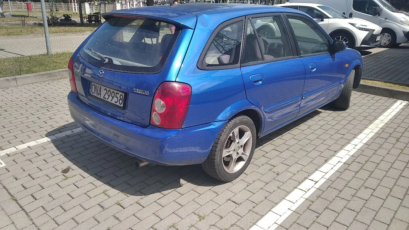 Niebieski samochód marki Mazda z nadwoziem typu hatchback zaparkowany na kostce brukowej. Widoczna jest tylna część pojazdu z tablicą rejestracyjną o numerze ONA 29958 oraz napisem Mazda. W tle widoczne są inne zaparkowane samochody i fragmenty infrastruktury miejskiej.