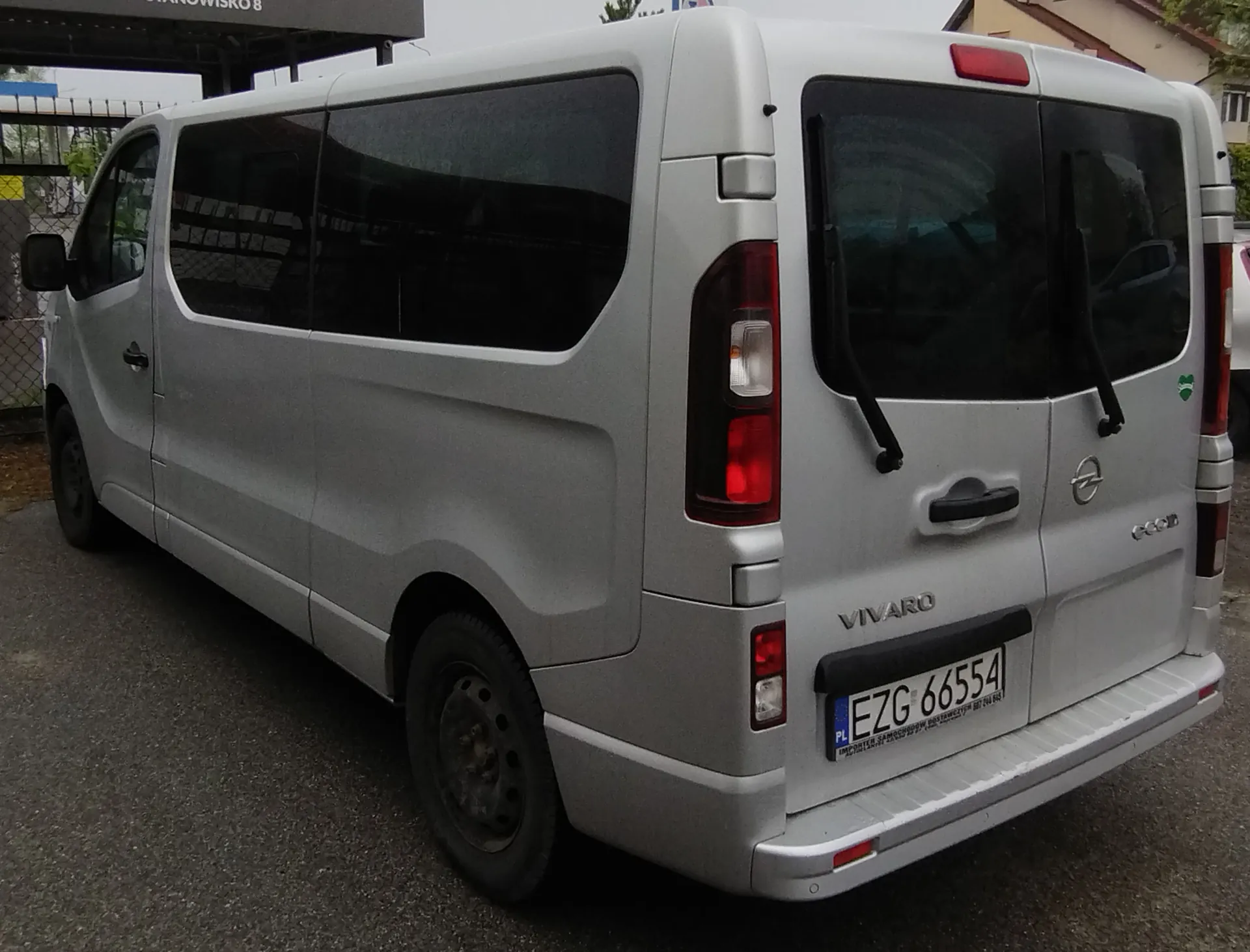 Srebrny samochód dostawczy marki Opel Vivaro zaparkowany na asfaltowej nawierzchni. Widok z tyłu i z boku pojazdu. Samochód ma przyciemniane szyby, czarne felgi oraz polską tablicę rejestracyjną o numerze EZG 66554. Na tylnych drzwiach widnieje napis VIVARO oraz logo marki Opel.