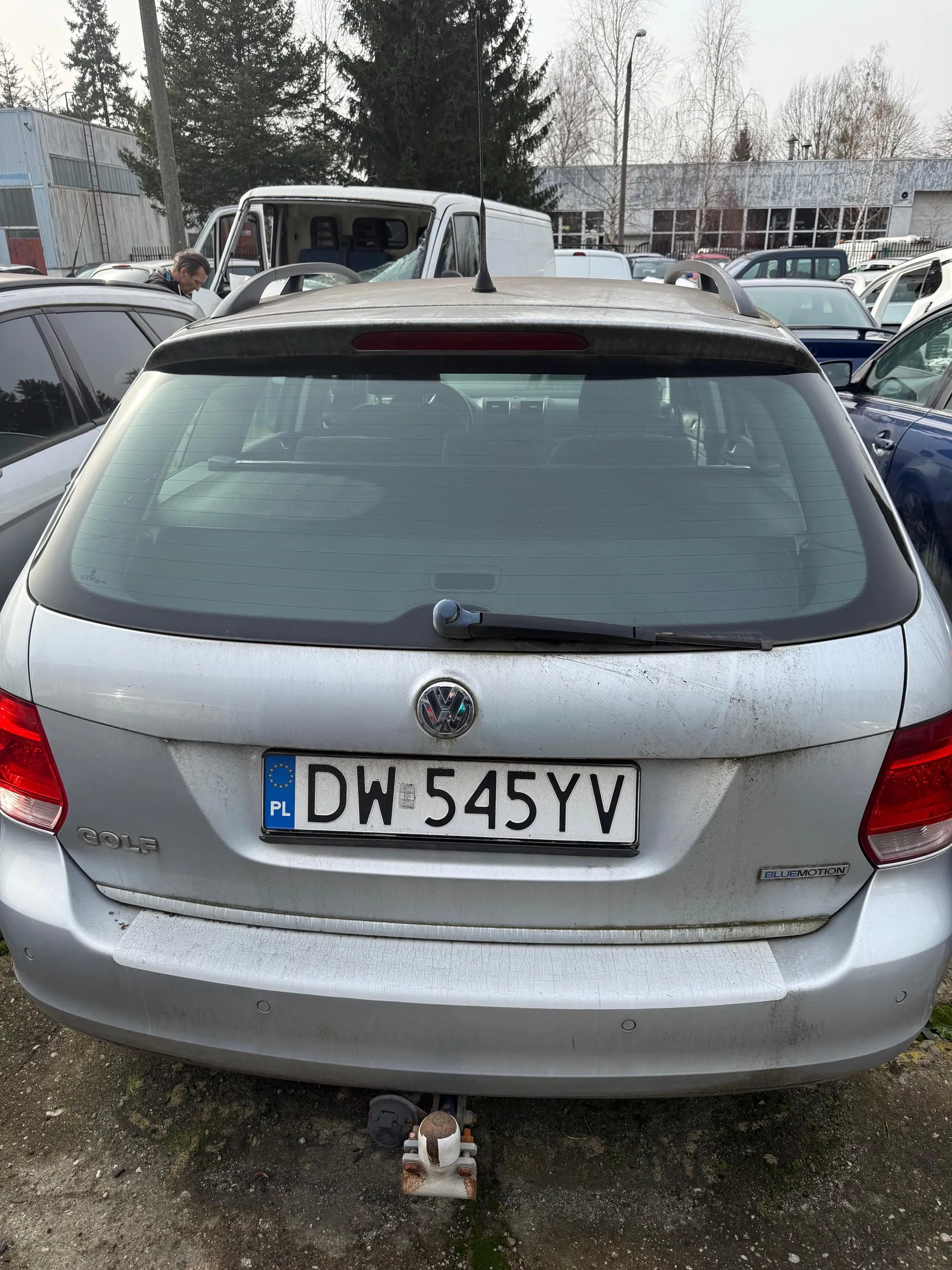 Tył srebrnego samochodu Volkswagen Golf z widocznym logo marki, tablicą rejestracyjną PL DW 545YV oraz napisem BLUEMOTION po prawej stronie. Na lewej stronie widoczny napis GOLF. Samochód stoi na podwórku z innymi pojazdami, w tle budynki i drzewa. Na dnie kadru widoczny holownik. Tablica rejestracyjna i tekst na samochodzie są poziomo ustawione.