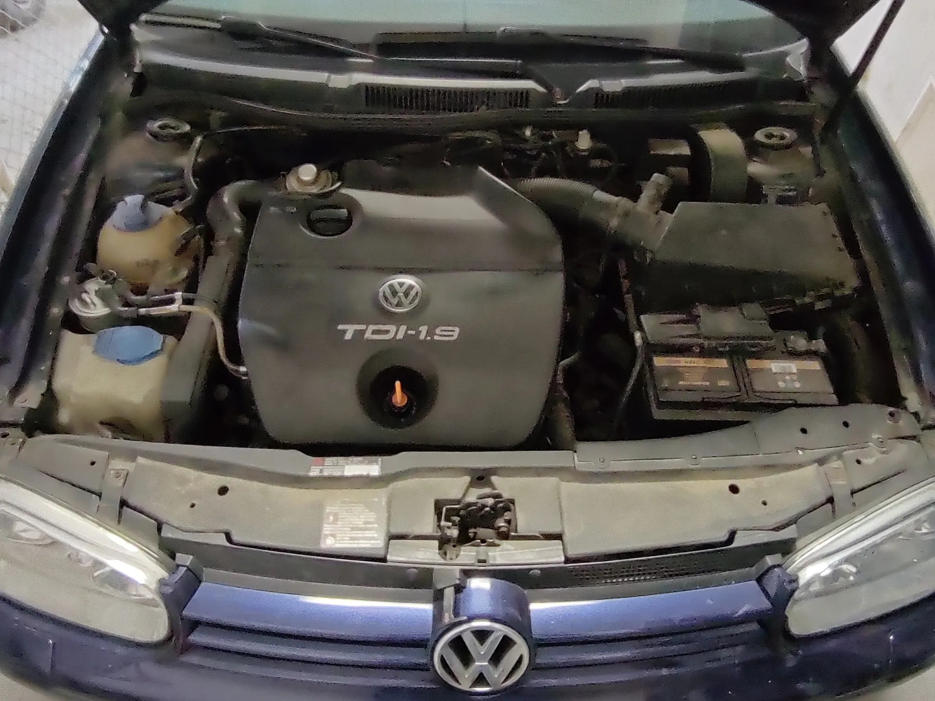 Widok otwartej tylnej części silnika samochodu marki Volkswagen. Na pokrywie silnika widoczny jest napis "TDI-1.9" oraz logo VW. Po lewej stronie znajduje się zbiornik chłodniczy z niebieską pokrywką, po prawej bateria. Na dole obrazu widoczna jest część pasa przedniego z logo VW na atrapie chłodnicy oraz reflektory. Silnik i elementy pod maską są ułożone symetrycznie, a tekst na pokrywie silnika jest czytelny i poziomy.