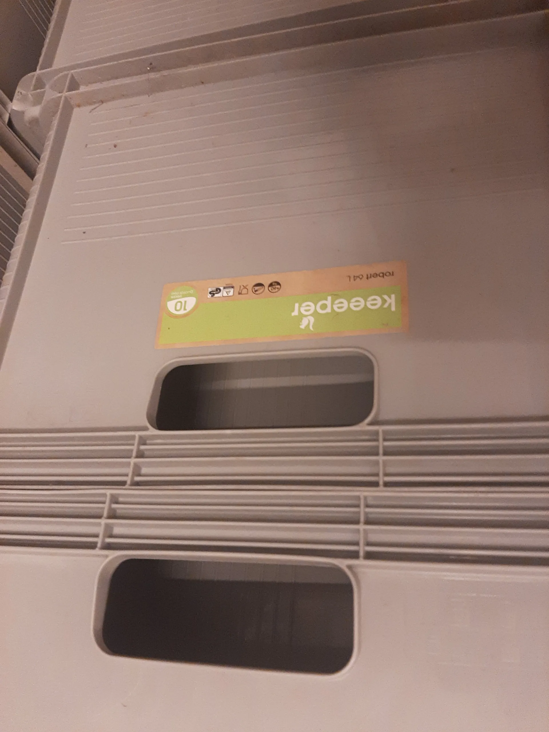 Biały plastikowy pojemnik z dwoma prostokątnymi otworami uchwytnymi po lewej stronie. Na prawo od otworów znajduje się etykieta w kolorach zielonym i brązowym z pionowo umieszczonym napisem 'keeper', cyfrą '10' w okręgu, ikonami oraz napisami 'robert' i '64 L'.