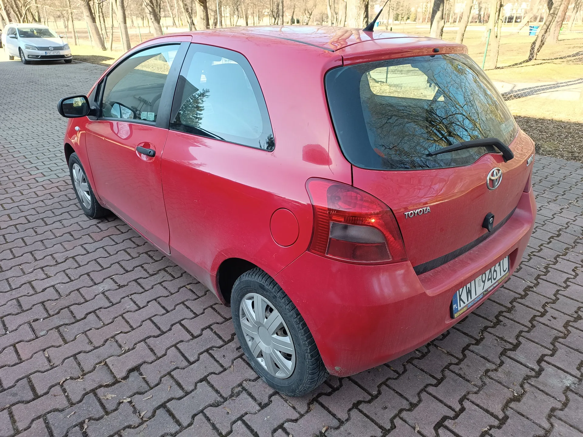Czerwony samochód Toyota Yaris trzydrzwiowy zaparkowany na brukowanej powierzchni. Widoczny tył pojazdu z logo Toyota, tablicą rejestracyjną KWI 9461C oraz czerwonymi światłami tylnymi. W tle biały samochód, drzewa bez liści i ogrodzenie. Dzień słoneczny, cień pada na bruk.