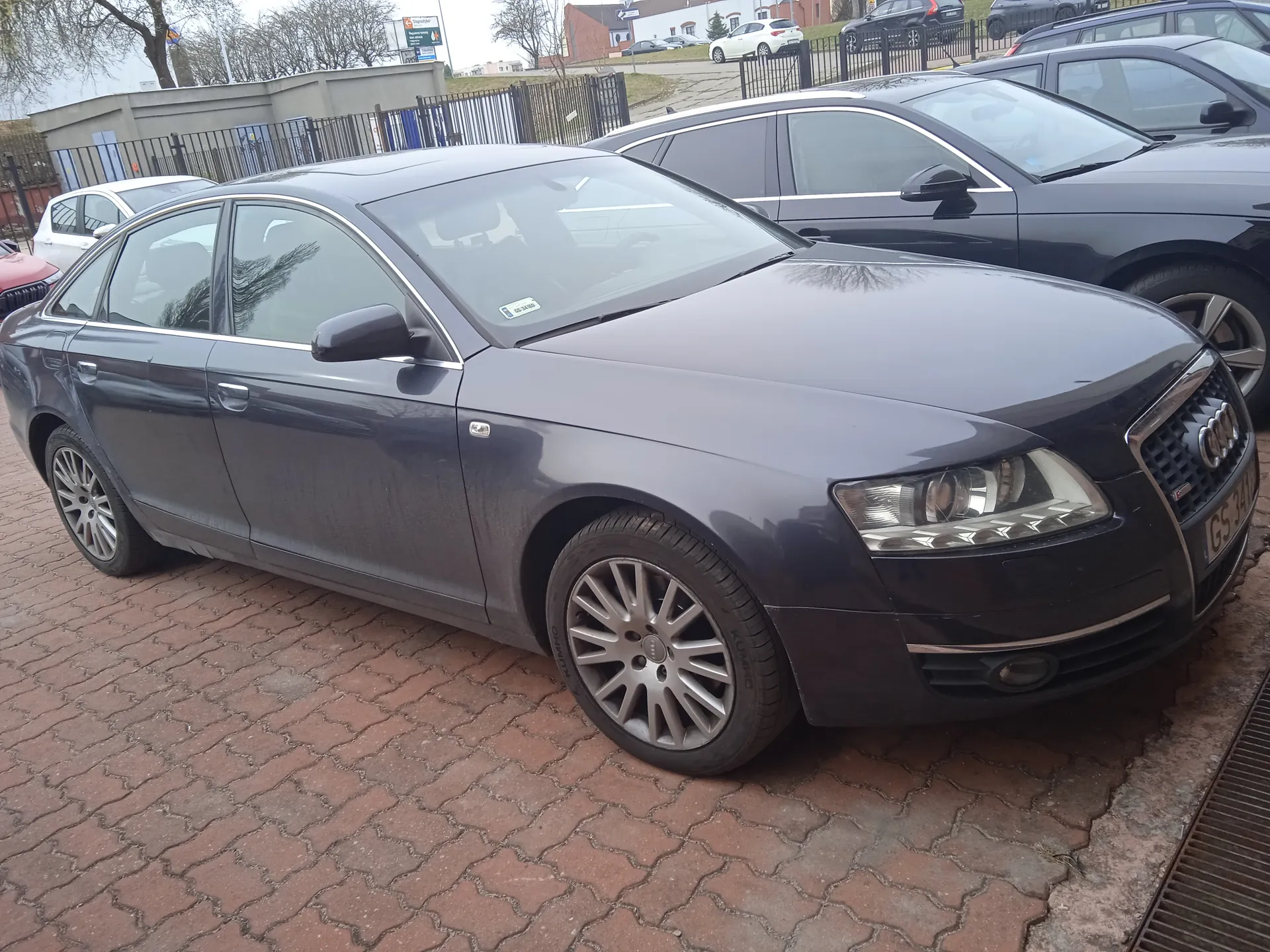 Ciemnoszary samochód Audi sedan zaparkowany na ceglanym placu parkingowym. W tle widoczne są inne pojazdy, w tym czarny SUV i biały samochód, ogrodzenie metalowe, budynki oraz drzewa. Na tablicy rejestracyjnej widoczny jest napis GS 3410. Niebo jest zachmurzone.