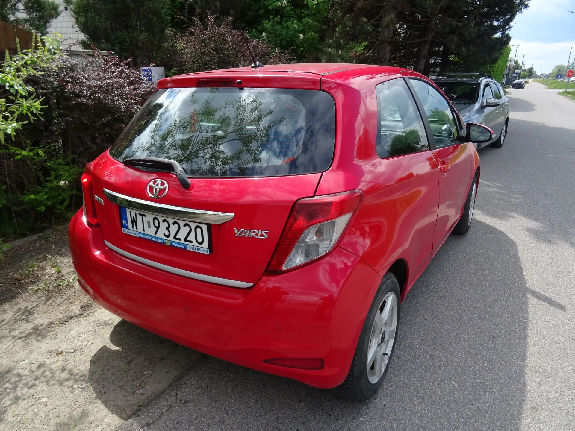 Czerwony samochód marki Toyota Varis zaparkowany przy krawędzi asfaltowej drogi. Widoczny jest tył pojazdu z tablicą rejestracyjną o numerze WT 93220. W tle widać zielone krzewy, drzewa oraz inny, srebrny samochód zaparkowany dalej na drodze.