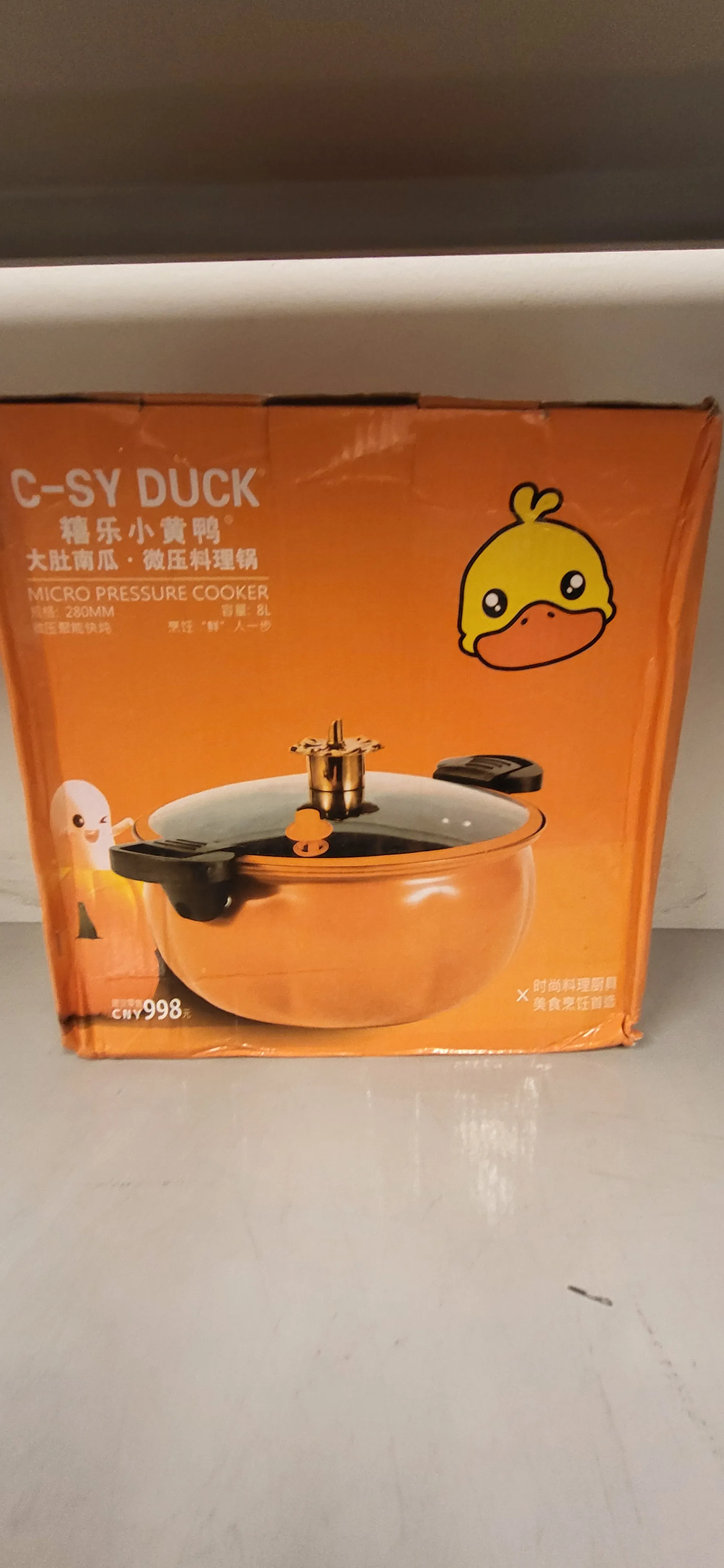 Pomarańczowe pudełko produktu z żółtym kaczątkiem w lewym górnym rogu. Na pudełku widoczny tekst "C-SY DUCK", chińskie znaki, napis "MICRO PRESSURE COOKER" oraz cena "CNY 998". W centralnej części znajduje się zdjęcie pomarańczowego garnka ciśnieniowego z czarnymi uchwytami i białą pokrywką. Tło stanowi biała powierzchnia.