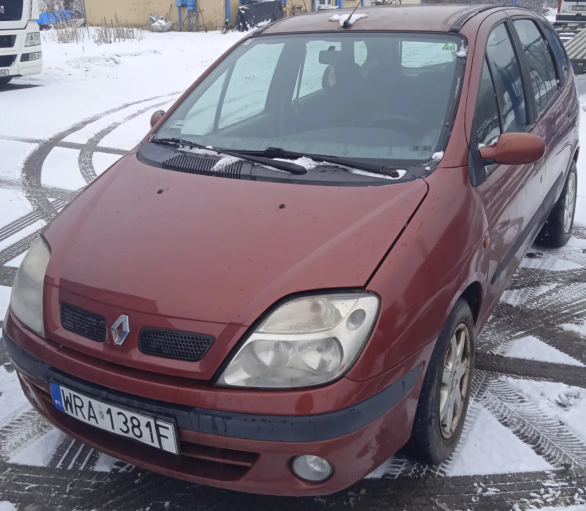 Czerwony samochód marki Renault z tablicą rejestracyjną WRA 1381F, zaparkowany na śnieżnej nawierzchni z widocznymi śladami opon. Na masce i przedniej szybie leży niewielka ilość śniegu. W tle biały pojazd ciężarowy oraz budynki przemysłowe. Na tablicy rejestracyjnej widoczny jest niebieski pas z napisem PL po lewej stronie.