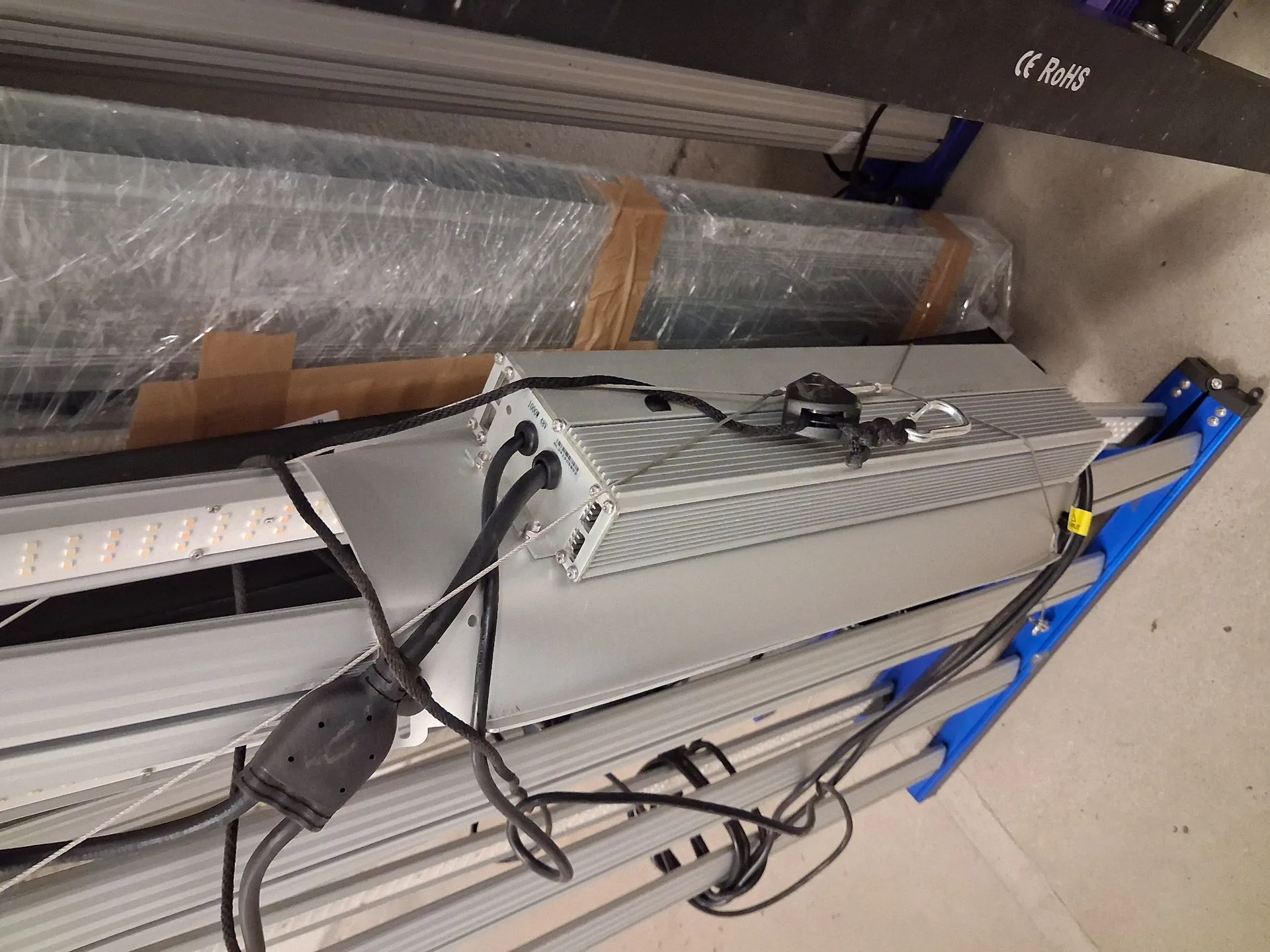 Zdjęcie przedstawia srebrny, aluminiowy radiator z zamontowanym zasilaczem i okablowaniem, leżący na szarych profilach aluminiowych. Obok znajduje się taśma LED. W tle widoczny jest przedmiot zapakowany w folię bąbelkową oraz niebieski element konstrukcyjny. W górnym prawym rogu widnieją oznaczenia 'CE RoHS'.