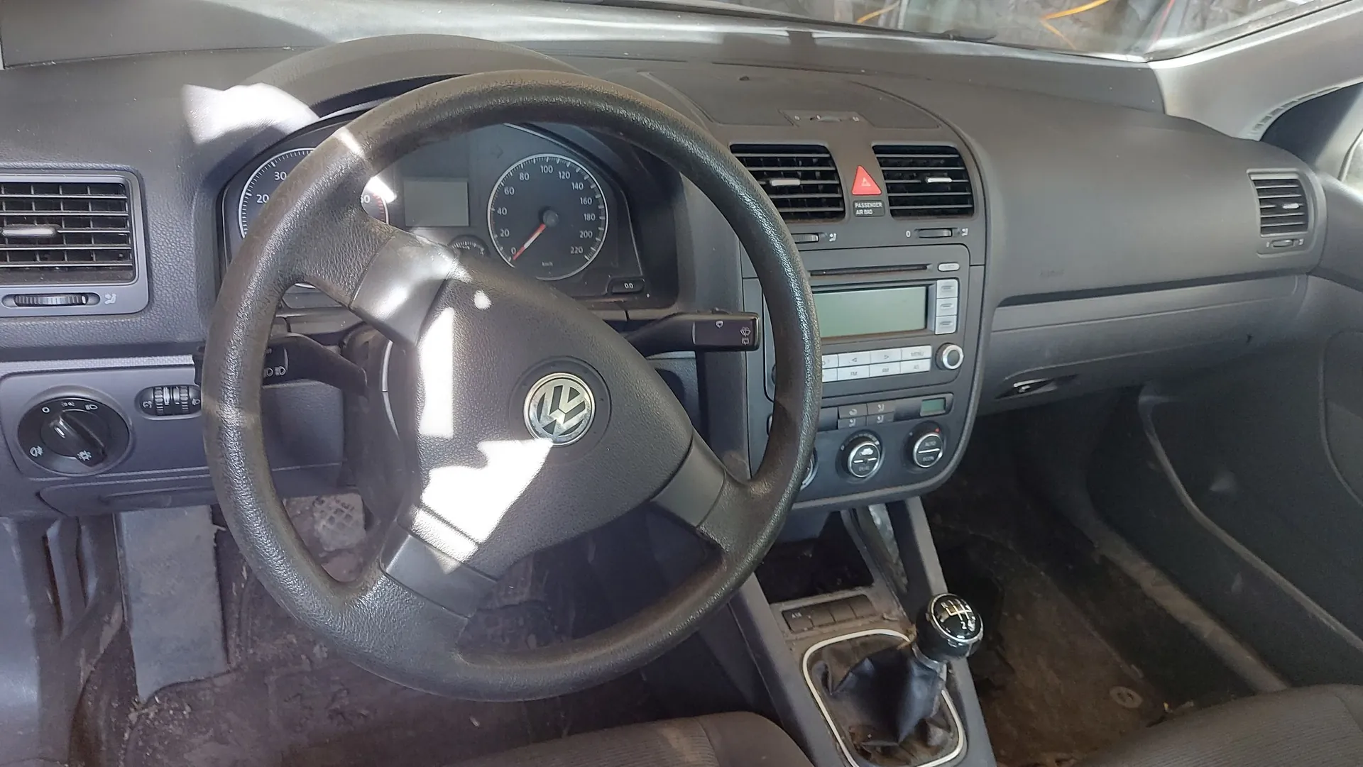Wnętrze samochodu marki Volkswagen widziane z perspektywy kierowcy. Widoczna jest czarna kierownica z logo VW w centrum, deska rozdzielcza z zegarami (prędkościomierzem i obrotomierzem), centralna konsola z radiem i pokrętłami klimatyzacji oraz lewarek zmiany biegów. Na kierownicę i deskę rozdzielczą pada jasne światło słoneczne.