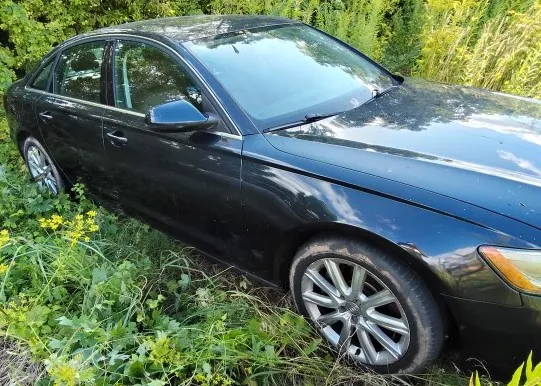 Czarny samochód osobowy typu sedan zaparkowany w wysokiej, zielonej trawie i roślinności. Pojazd jest widoczny z perspektywy przedniego lewego boku, z widocznym przednim kołem z aluminiową felgą oraz przednią lampą.