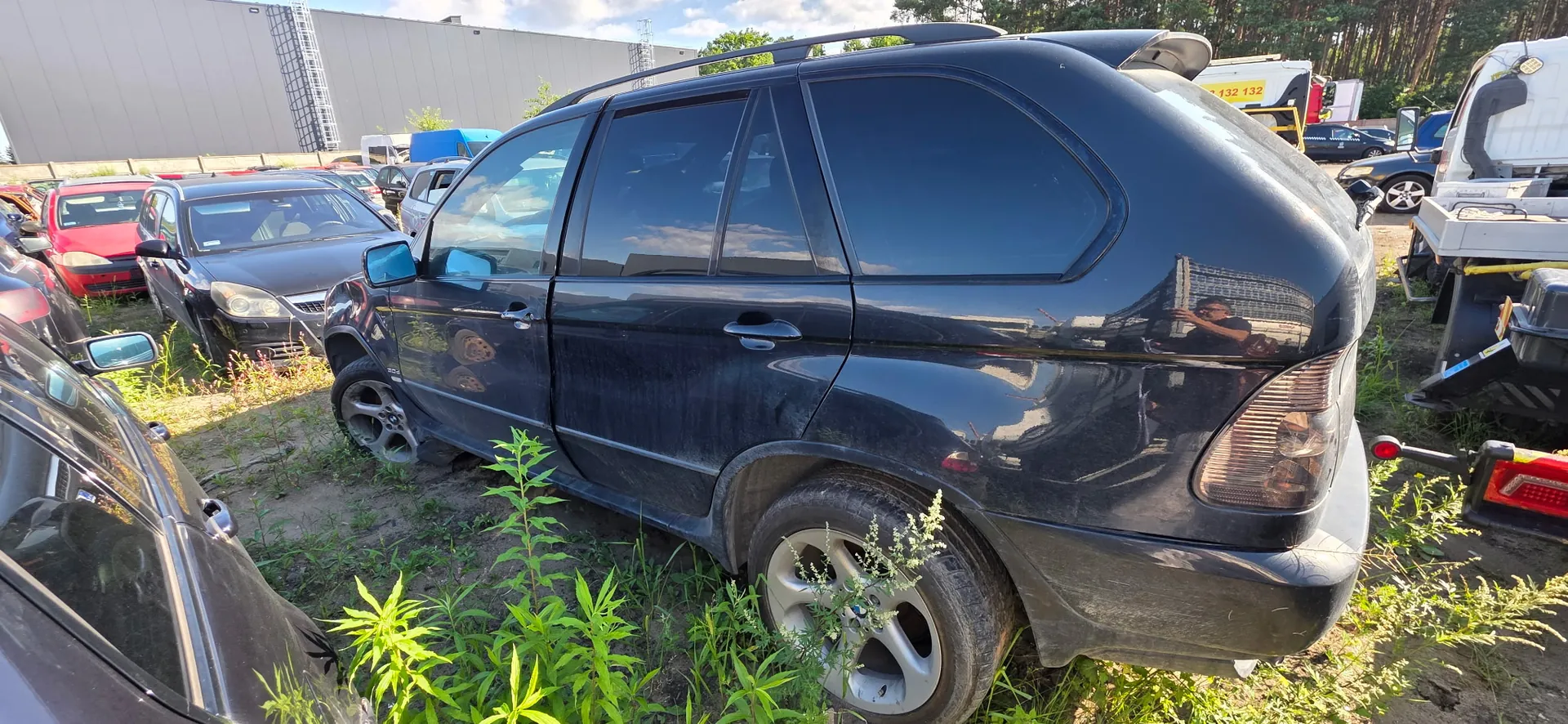 Czarny samochód typu SUV zaparkowany na trawiastym placu wśród innych pojazdów. Auto jest zakurzone, widoczny jest profil boczny i tył pojazdu. W tle znajdują się inne samochody, budynek przemysłowy oraz pas drzew.