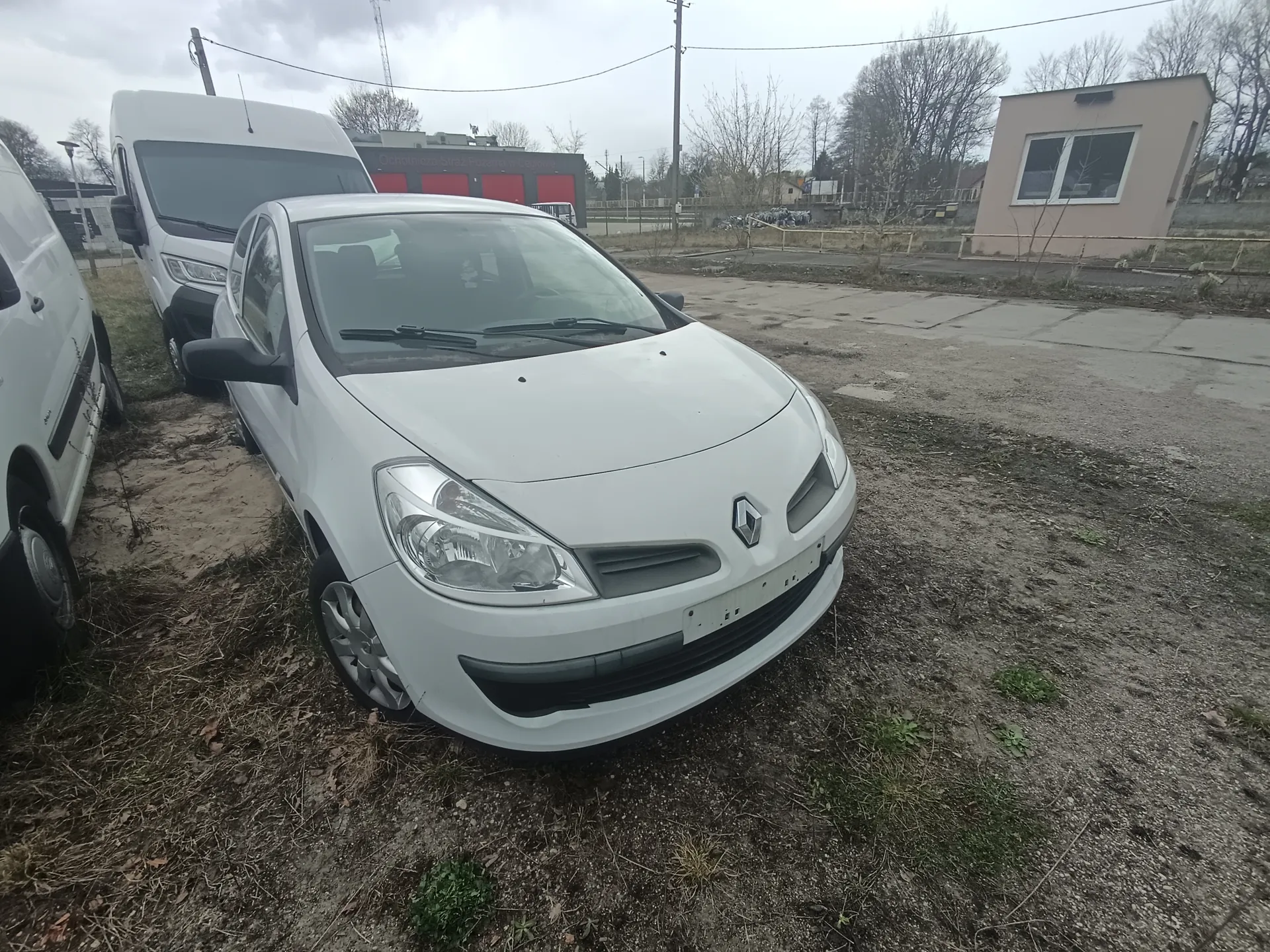 Trzy białe pojazdy – samochód osobowy marki Renault i dwa furgony – zaparkowane na terenie z żwirem i suchą trawą. W tle budynki, w tym czerwone drzwi, mały beżowy budynek z oknami, drzewa bez liści, słupy energetyczne oraz pochmurne niebo.