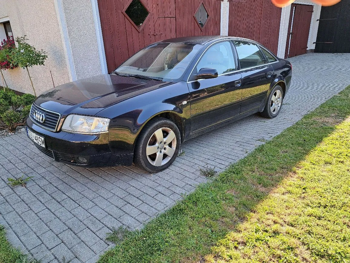 Czarny samochód marki Audi zaparkowany na kostce brukowej przed budynkiem z czerwonymi drzwiami garażowymi. Auto jest ustawione pod kątem, widoczny jest przód i prawy bok pojazdu.