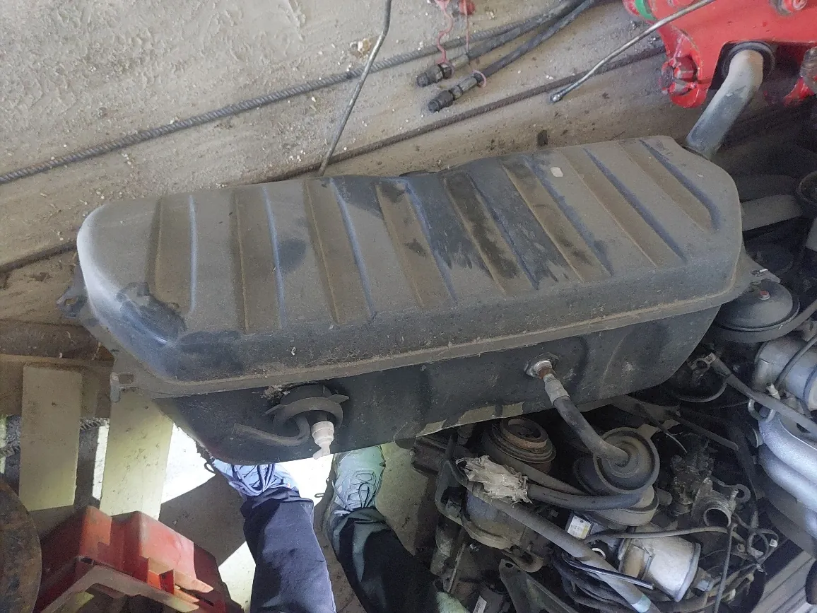 Zdjęcie przedstawiające czarną, plastikową obudowę filtra powietrza w komorze silnika samochodu. Obudowa jest zakurzona i ma żebrowaną strukturę na górze. W tle widoczne są inne elementy silnika, w tym paski i koła pasowe. Obraz jest zorientowany pionowo, podczas gdy komora silnika znajduje się w pozycji poziomej.