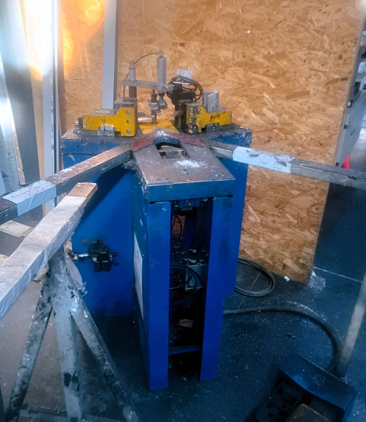 Niebieska maszyna przemysłowa z metalowymi prowadnicami i mechanizmem zaciskowym na górze. Urządzenie znajduje się w warsztacie na tle płyty OSB. Na górze widoczne są żółte elementy mocujące oraz siłownik pneumatyczny.