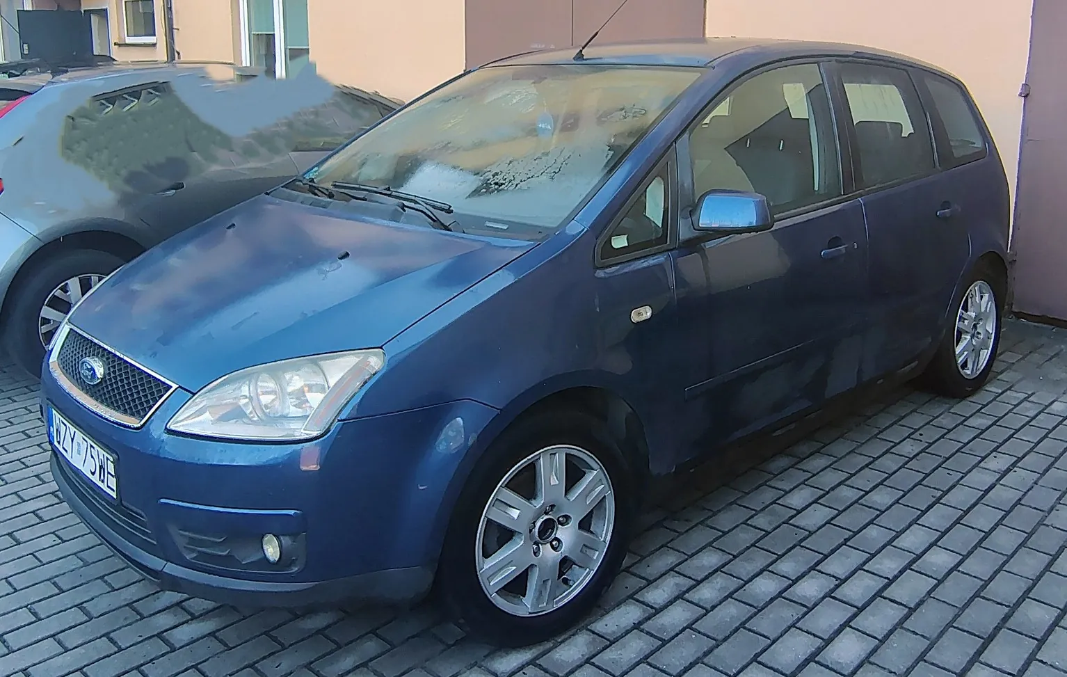 Niebieski samochód osobowy marki Ford w nadwoziu typu kombi/minivan, zaparkowany na kostce brukowej. Widoczna jest przednia część pojazdu z lewej strony, tablica rejestracyjna o numerze WZY 75WE oraz fragment innego, srebrnego samochodu w tle po lewej stronie.