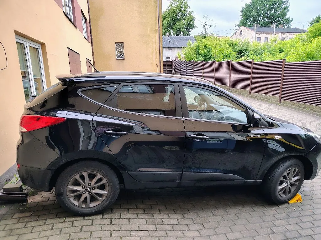 Czarny samochód typu crossover zaparkowany bokiem na kostce brukowej przed żółtym budynkiem. W tle widoczny jest brązowy płot z poziomych listew oraz zielone drzewa i krzewy.