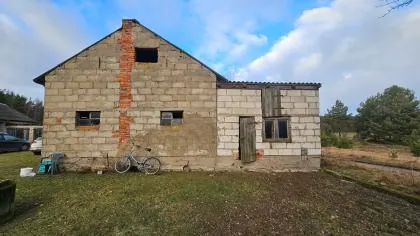 Zdjęcie starego, częściowo niedokończonego budynku z pustaków betonowych. Budynek składa się z wyższej części z dwuspadowym dachem i niższej dobudówki po prawej stronie. Ściany są szare, z widocznym pionowym pasem z czerwonej cegły. W budynku znajdują się małe, prostokątne okna oraz jedne drewniane drzwi w niższej części. Przed budynkiem, na trawiastym podłożu, stoi jasnoniebieski rower. W tle widoczne są drzewa i niebo z białymi chmurami.