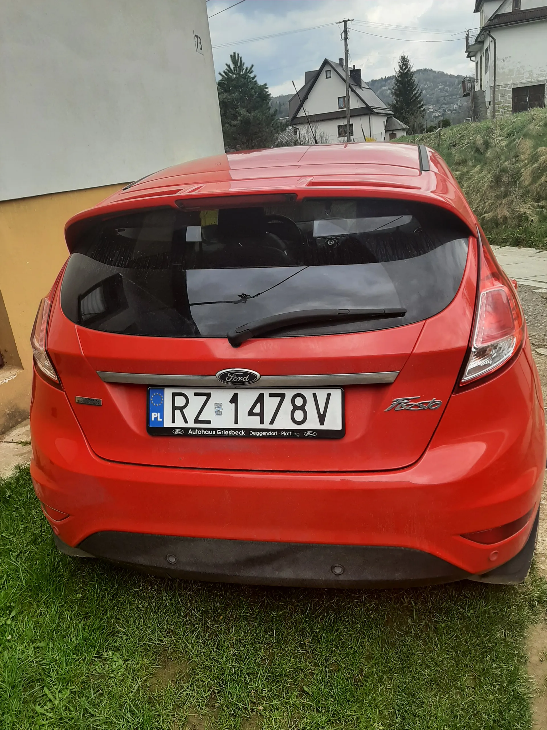 Czerwony samochód Ford Fiesta widziany od tyłu. Tablica rejestracyjna z białym tłem i czarnymi literami RZ 1478V oraz niebieskim pasem z napisem PL. Na ramce tablicy widoczny napis Autohaus Griesbeck Deggendorf - Plattling. W tle trawa, domy i niebo.